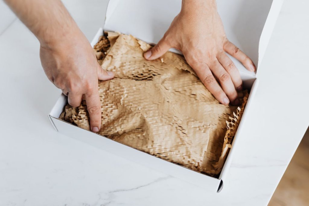 pexels-photo-4464482 From above crop adult male packing ordered product or gift in white cardboard box with craft paper for safety postal delivery at white marble table